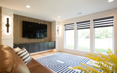 Modern basement lounge area featuring a large wall-mounted TV, striped area rug, sleek cabinetry, stylish lighting, and abundant natural light from large windows, designed for both relaxation and entertainment.