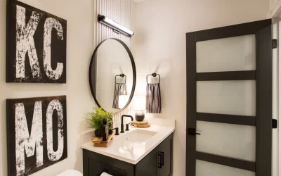 Modern bathroom design featuring a stylish sink area with matte cabinetry, circular mirror, and contemporary lighting, complemented by Kansas City-themed wall art and a glass door.